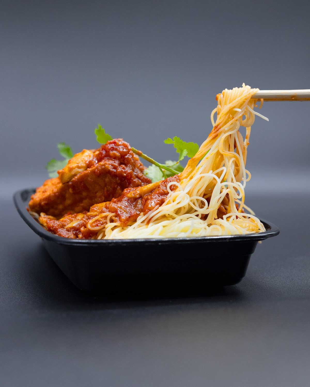Chopsticks lifting noodles beside grilled meat in a black takeaway tray